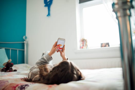 Little girl is lying on her bed, playing a game on her smartphone.