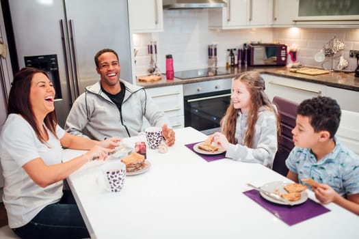 Family of four are enjoying breakfast together at home. They are all laughing while they have toast and jam.