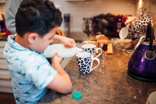 Little boy is pouring milk in to a mug while making tea with his father at home.