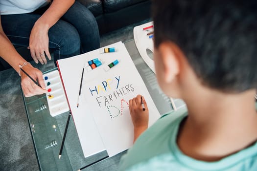 Little boy and his mother are making a Father's Day card at home using paints and crayons.