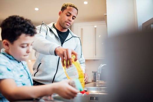 Little boy and his father are washing the dishes together at home. They are adding washing-up liquid to the sink.