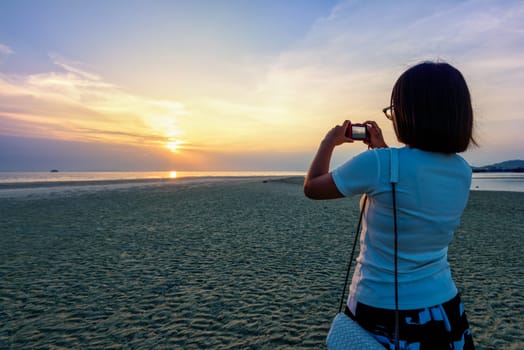 Asian woman tourist taking photo on the beach and beautiful natural landscape of colorful sky and sea during a sunset at Nathon Sunset Viewpoint in Samui island, Surat Thani, Thailand