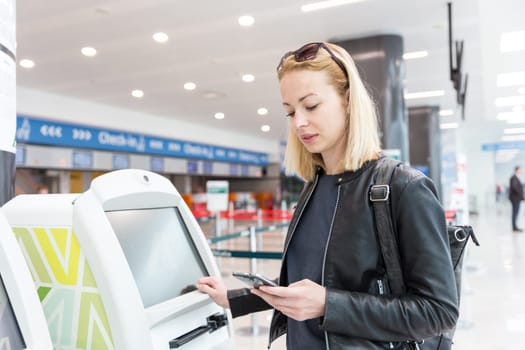 Casual caucasian woman using smart phone application and check-in machine at the airport getting the boarding pass. Modern technology on airport.