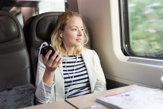 Businesswoman communicating on cellphone using headphone set while traveling by train in business class seat.