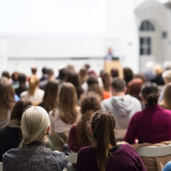 Female speaker giving presentation in lecture hall at university workshop. Audience in conference hall. Rear view of unrecognized participant in audience. Scientific conference event.