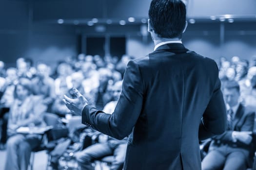 Speaker giving a talk on corporate business conference. Unrecognizable people in audience at conference hall. Business and Entrepreneurship event. Blue toned grayscale image.
