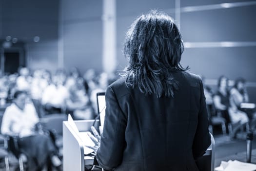 Female speaker giving a talk on corporate business conference. Unrecognizable people in audience at conference hall. Business and Entrepreneurship event. Black and white, blue toned image.