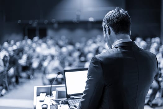 Speaker giving a talk on corporate business conference. Unrecognizable people in audience at conference hall. Business and Entrepreneurship event. Blue toned grayscale image.