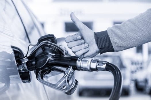 Petrol or gasoline being pumped into a motor vehicle car. Closeup of mans hand showing thumb up gesture, pumping gasoline fuel in car at gas station. Greyscale blue toned image.