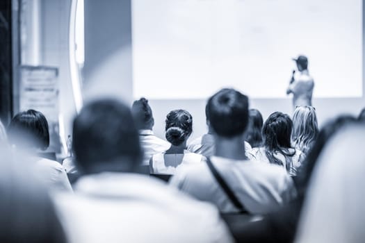 Speaker giving a talk in conference hall at business event. Focus on unrecognizable people in audience. Business and Entrepreneurship concept. Blue toned greyscale image.