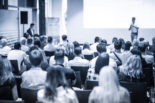Speaker giving a talk in conference hall at business event. Focus on unrecognizable people in audience. Business and Entrepreneurship concept. Blue toned greyscale image.