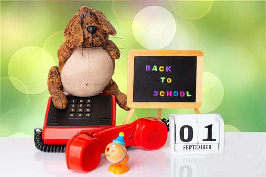 Colorful equipment with funny dog and toy clown on white table against light green colored bokeh background. Back to school written with colorful letters on blackboard. A wooden calendar shows the 01 september if starts school. Back to school.