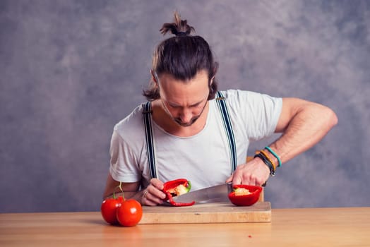 young bearded man cutting sweet pepper in front of gray background