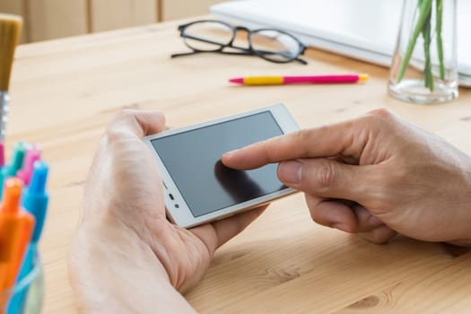 Businessman touching screen of smartphone close up. Touching smartphone in home office on wood table with pen and glasses and laptop