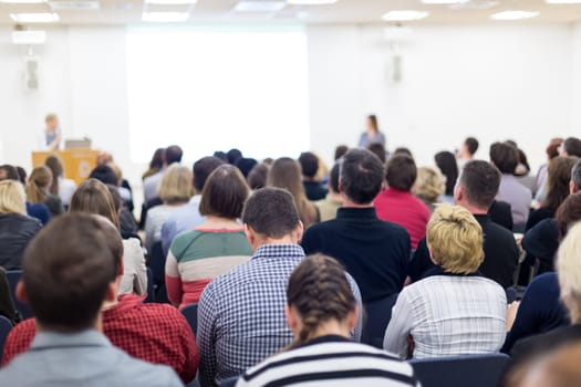 Business and entrepreneurship symposium. Female speaker giving a talk at business meeting. Audience in conference hall. Rear view of unrecognized participant in audience. Copy space on whitescreen.