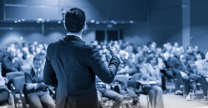 Speaker giving a talk on corporate business conference. Unrecognizable people in audience at conference hall. Business and Entrepreneurship event. Blue toned grayscale image.