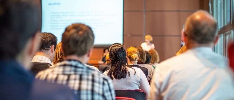 Business and entrepreneurship symposium. Female speaker giving a talk at business meeting. Audience in conference hall. Rear view of unrecognized participant in audience.
