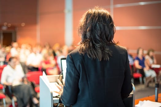 Female speaker giving a talk on corporate business conference. Unrecognizable people in audience at conference hall. Business and Entrepreneurship event.