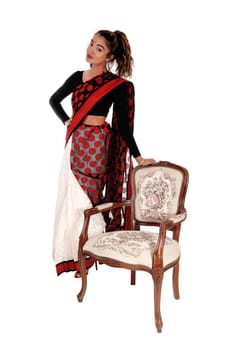A lovely young East Indian woman in a colorful saree standing beside an.old fashion armchair, isolated for white background.