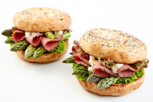 Two gourmet asparagus burgers with fresh green tips and sliced parma ham topped with mayo served on buns over white with copy space