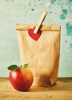 School lunch in a brown paper packet pegged with a heart alongside a fresh ripe healthy red apple on a wooden table