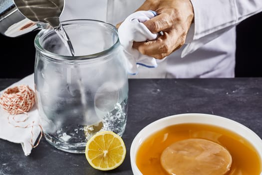 Man sterilising a glass jar pouring boiling water from a jug as he prepares a container to make homemade kombucha with the scoby starter or fungus mat in the foreground