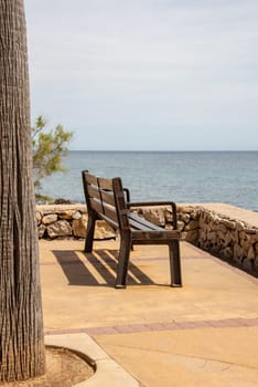 A bench and a single tree on the promenade in the holiday resort Cala Millor with a beautiful view of Mittelmerr on the Spanish Balearic island Mallorca in front of a radiant blue sky
