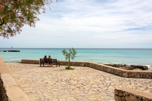 View of the turquoise Mediterranean Sea and the beach promenade with a bench in the holiday resort Cala Millor on the Spanish Balearic island Mallorca in front of a bright blue sky