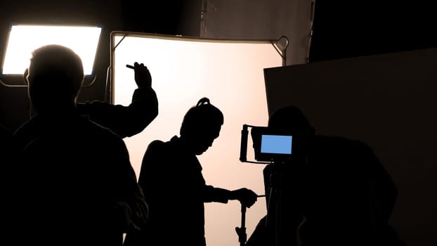 Video production behind the scenes which film crew team in silhouette shooting or recording tv movie commercial with professional equipment such as high definition 4k camera with monitor and tripod or gimbal rig in big studio set.