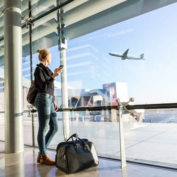 Young casual female traveler at airport, holding smart phone device, looking through the airport gate windows at planes on airport runway.