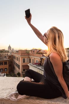 F ashinable female tourist with a vintage hipster backpack taking selfie photo with her mobile phone of Piazza di Spagna, landmark square with Spanish steps in Rome, Italy at sunset.
