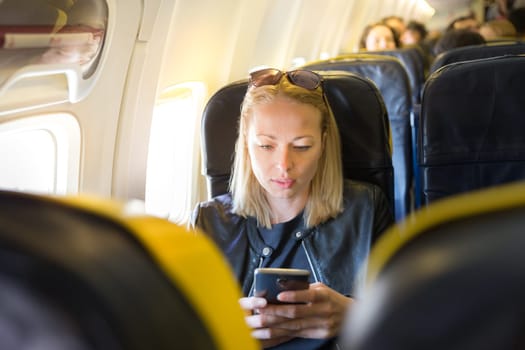Woman using mobile phone as entertainment on airplane during commercial flight. Female traveler seated in passanger cabin. Sun shining trough airplane window.