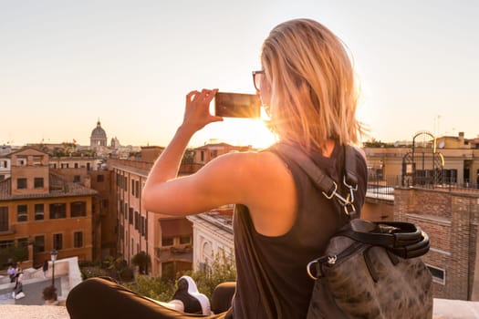 Female tourist with a fashinable vintage hipster backpack taking mobile phone photo of Piazza di Spagna, landmark square with Spanish steps in Rome, Italy at sunset.