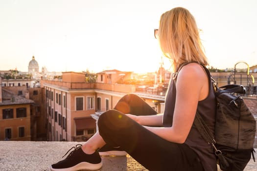Fashinable female tourist with vintage hipster backpack admiring the beautiful view of at Piazza di Spagna, landmark square with Spanish steps in Rome, Italy at sunset.