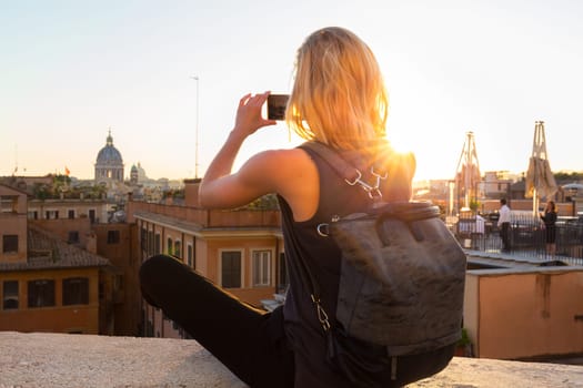 Female tourist with a fashinable vintage hipster backpack taking mobile phone photo of Piazza di Spagna, landmark square with Spanish steps in Rome, Italy at sunset.