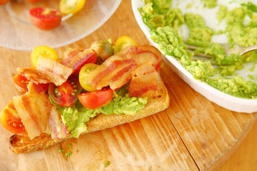 Toast, mashed avocados, cherry tomatoes and bacon on a cutting board