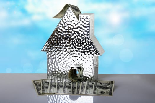 A metallic house with one hundred dollar bills on table with bright blue background. Space for text display montage. Concept of money and earnings.