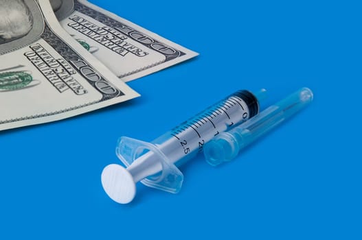 A syringe, and one hundred dollar bills on a blue background. Side view.