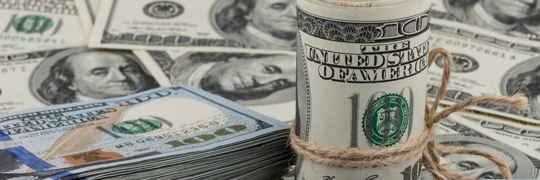 A roll of one hundred dollar bills tied with a rope against the backdrop of scattered dollars.
