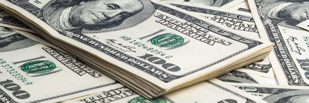 A pack of one hundred dollar bills in the background of scattered dollars. View at an angle