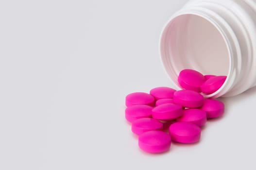 Brightly pink tablets scatter from a white bottle. Close-up.