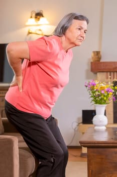 Mature woman suffering from backache at home. Massaging lower back with hand, feeling exhausted, standing in living room.