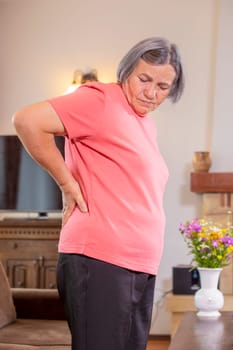 Mature woman suffering from backache at home. Massaging lower back with hand, feeling exhausted, standing in living room.