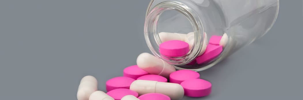 White pills and pink tablets are scattered from a glass bottle on a gray background.