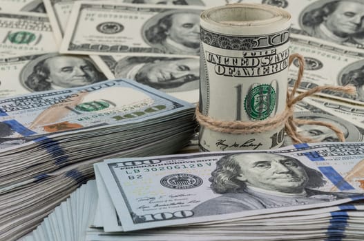 A roll of one hundred dollar bills tied with a rope against the backdrop of scattered dollars. Side view.