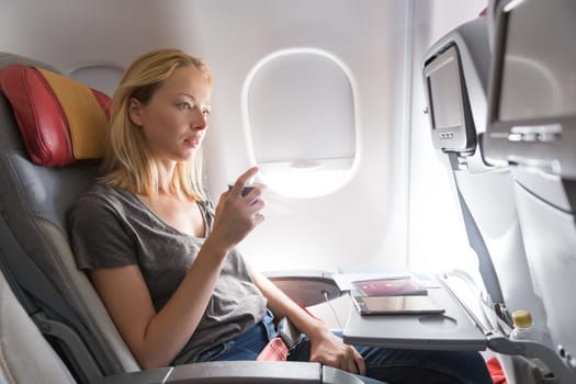 Woman on commercial passengers airplane during flight. Female traveler seated in passanger cabin drinking coffee. Sun shining trough airplane window.