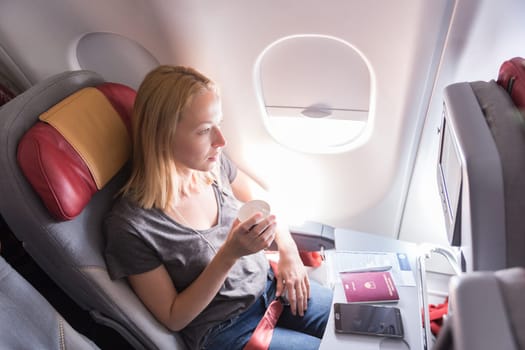 Woman on commercial passengers airplane during flight. Female traveler seated in passanger cabin drinking coffee. Sun shining trough airplane window.