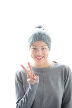 Close up portrait of cheerful caucasian blond woman, gesturing peace sign and smiles, wearing casual clothes and gray hat against white background. Copy space.