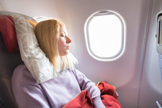 Tired blonde casual caucasian woman sleeping on seat while traveling by airplane on long distance transatlantic flight. Commercial transportation by planes.