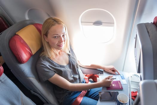 Relaxed casual woman flying on commercial passengers airplane, filling in immigration form, drinking coffee, listening to music, smiling, looking at camera.